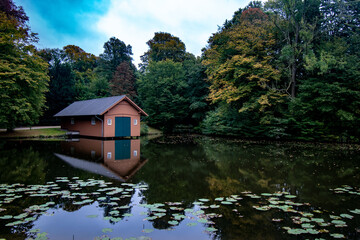 Early autumn view of a wooden cabin by reflective pond water with lily pads in Burger Park, Germany
