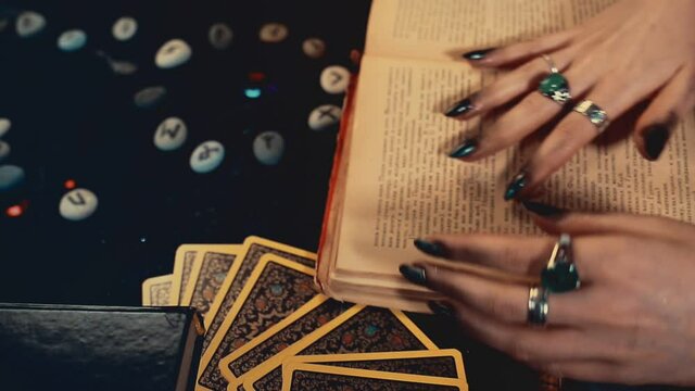 Divination and magic. A fortune teller leafs through a book of spells. Movement of the camera from top to bottom, from the face to the table with books, Tarot cards and runes.