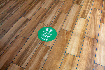 sign on wooden floor. please queue here sign. safe distancing, covid 19, coronavirus.