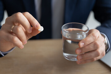 Close up young businessman in formal wear with pill and glass of water thinking of taking painkiller or antibiotic aspirin feeling unwell in office, millennial worker employee holding daily vitamins