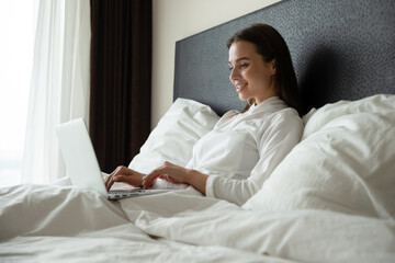 No need in leaving bed. Happy smiling young female programmer remote employee freelancer using laptop while sitting in comfortable bed at home hotel looking at screen chatting online surfing internet