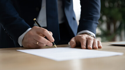 Close up young businessman standing near table with pen in hands, ready signing profitable offer agreement after checking contract terms of conditions, executive manager involved in legal paperwork. - obrazy, fototapety, plakaty
