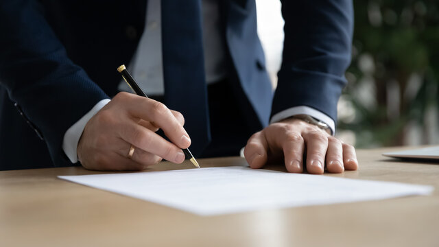 Close up young businessman standing near table with pen in hands, ready signing profitable offer agreement after checking contract terms of conditions, executive manager involved in legal paperwork.