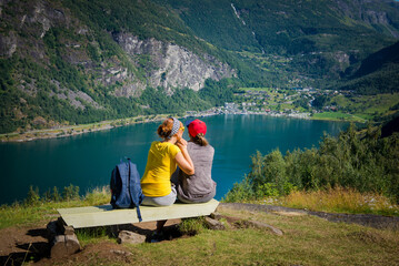 Fototapeta na wymiar mom and daughter sit on the bench with their backs. Norway, view from the mountain to the city of Geiranger and the fjord. Mom and daughter together.
