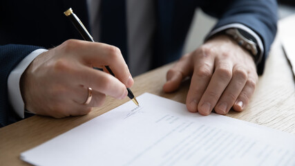 Close up young entrepreneur signing paper contract or business legal agreement, sitting at table. Focused male ceo executive manager putting signature on company financial or law document in office.