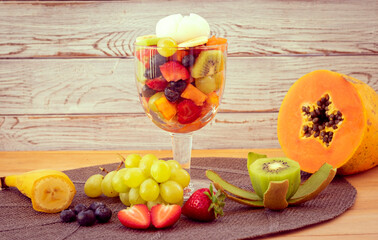 A glass cup with healthy fresh colorful fruit salad ready to eat with grapes, strawberry, papaya, banana, kiwi and blueberries. On top a white ice cream. Wooden background