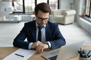 Focused serious young businessman in formal suit checking time on wristwatch, planning future...