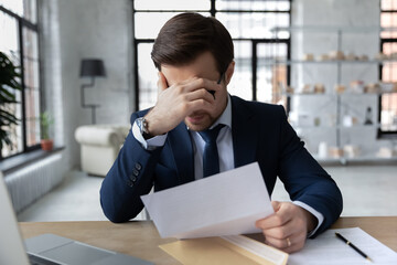 Frustrated 30s employee in formal wear holding paper dismissal notice, unhappy confused young...