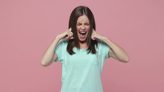 Dissatisfied young woman 20s years old in blue t-shirt posing isolated on pink background studio. People lifestyle concept. Hold hands crossed covering ears with fingers keeping eyes closed screaming