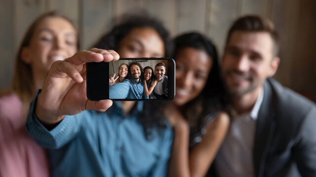 Taking group selfie. Blurred portrait of four diverse multiethnic millennial friends students coworkers hugging smiling and looking at camera showing spectator self picture making on smartphone webcam