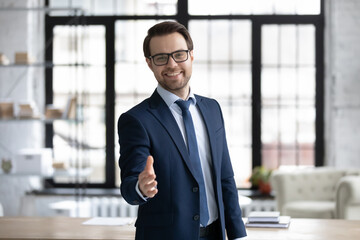 Happy young businessman in glasses looking at camera, reaching out hand making welcoming gesture or ready handshaking with partner or colleague, smiling banker proposing good deal to client.