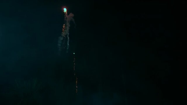 Red and green fireworks are displayed in the night sky illuminating the trees. The rockets leave a trail of smoke as each firework skyrocket explodes.