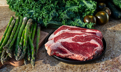 Raw meat, beef steak on black plate with kale, asparagus, broccolini, and tomatoes in the background.