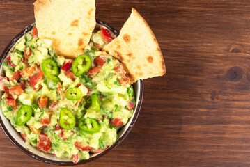 Homemade guacamole - traditional mexican appetizer of avacado, onion and tomato in a bowl with toasted tortilla pieces on a wooden background, copy space