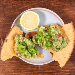 Homemade guacamole - a traditional Mexican appetizer of avacado, onion and tomato on toasted tortilla pieces in a plate with half a lime on a rustic wooden table