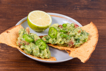 Homemade guacamole - a traditional Mexican appetizer of avacado, onion and tomato on toasted tortilla pieces in a plate with half a lime on a rustic wooden table