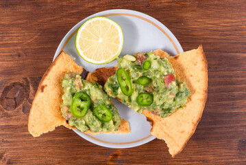 Homemade guacamole - a traditional Mexican appetizer of avacado, onion and tomato on toasted tortilla pieces in a plate with half a lime on a rustic wooden table