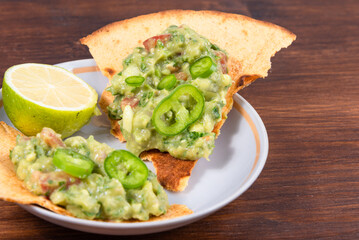 Homemade guacamole - a traditional Mexican appetizer of avacado, onion and tomato on toasted tortilla pieces in a plate with half a lime on a rustic wooden table