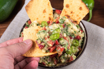 Homemade Guacamole - Hand holds a piece of toasted szakuca tortilla on top of a plate on a rustic wooden table
