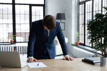 Frustrated young businessman in formal wear standing at office table with head down, feeling depressed after professional failure, company bankruptcy, money loss or unprofitable deal concluding. - obrazy, fototapety, plakaty