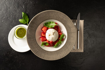 Mozzarella cheese with fresh tomato and basil salad. Served with pesto sauce. Veggie platter for restaurant menu served with cutlery on a black stone table.