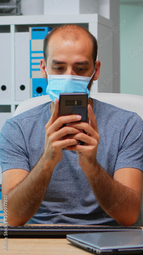 Poster office worker with protective mask using phone typing sitting on modern office during coronavirus. f