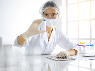 Professional female scientist in protective eyeglasses researching tube with reagents in sunny laboratory. Medicine and researching