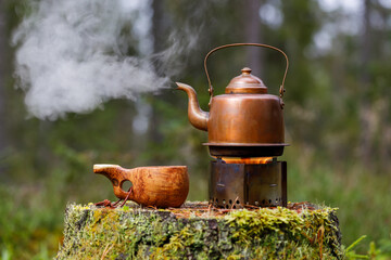 Vintage kettle on a tourist stove with a wooden cup on an old stump in the forest. © Mikhail