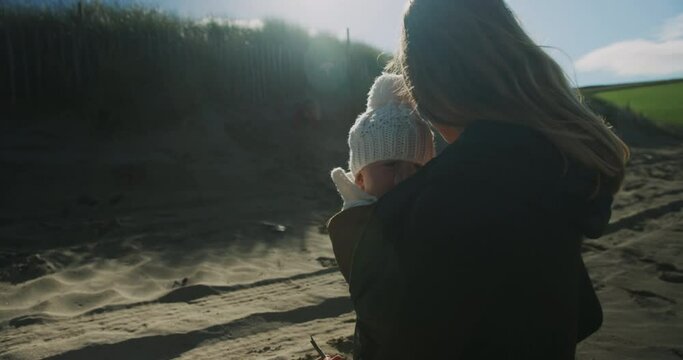 A young mother with her baby in a sling is watching her preschooler play on the beach in autumn
