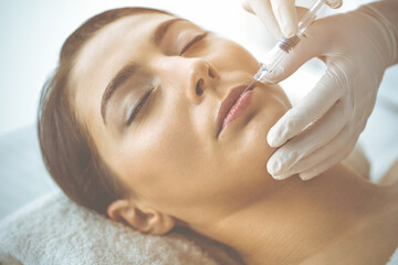 Naklejka premium Beautician doing beauty procedure with syringe to face of young brunette woman in sunny clinic. Cosmetic medicine and surgery, beauty injections