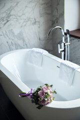 A delicate wedding bouquet of roses lies on the edge of a snow-white bathroom. Designer bathroom in the center of the bathroom. White artificial stone bathtub. Designer bathroom interior