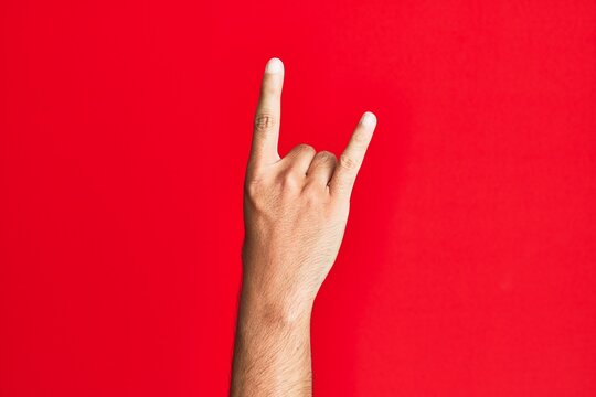 Arm of caucasian white young man over red isolated background gesturing rock and roll symbol, showing obscene horns gesture