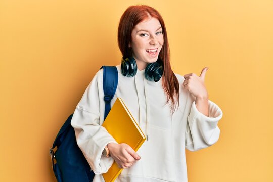 Young red head girl wearing student backpack and holding book pointing thumb up to the side smiling happy with open mouth