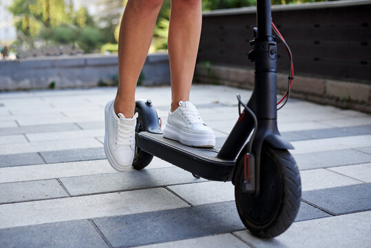 Close-up picture of woman's legs, riding on electric scooter in city center. Female, wearing white sneakers. Summer leisure activity. Traveler exploring the town, spending time outdoors.