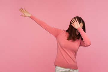 Positive popular young woman with brown hair in pink sweater making dab dance gesture, popular internet trends. Indoor studio shot isolated on pink background