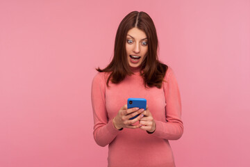 Excited brunette woman looking at smartphone display in her hands with big surprised eyes, receiving message about bonuses and discounts. Indoor studio shot isolated on pink background