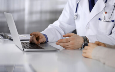 Fototapeta premium Unknown male doctor and patient woman discussing something while sitting in clinic and using laptop. Best medical service in hospital, medicine, pandemic stop