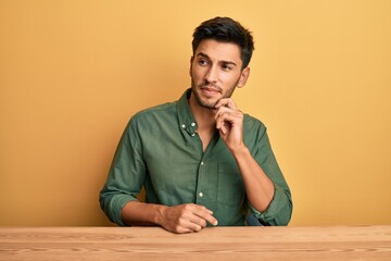 Young handsome man wearing casual clothes sitting on the table thinking concentrated about doubt...