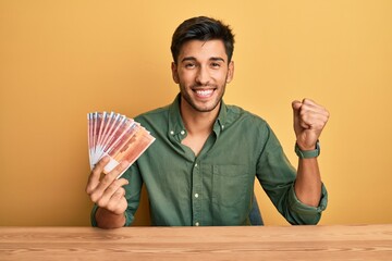 Young handsome man holding norwegian krone banknotes screaming proud, celebrating victory and...