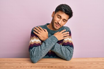 Young handsome man wearing casual winter sweater sitting on the table hugging oneself happy and...