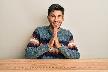 Fototapeta premium Young handsome man wearing casual sweater sitting on the table praying with hands together asking for forgiveness smiling confident.