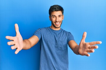 Young handsome man wearing casual tshirt over blue background looking at the camera smiling with open arms for hug. cheerful expression embracing happiness.