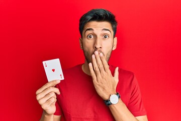 Young handsome man holding ace poker card covering mouth with hand, shocked and afraid for mistake....