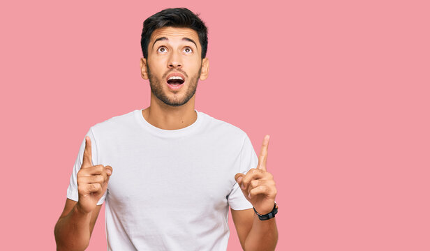 Young handsome man wearing casual white tshirt amazed and surprised looking up and pointing with fingers and raised arms.