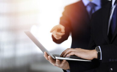 Business people discussing a presentation at meeting, standing in a sunny modern office. Unknown businessman with a colleague search for some information at the laptop, lawyers at negotiation