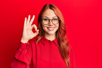 Young beautiful redhead woman wearing casual clothes and glasses over red background smiling positive doing ok sign with hand and fingers. successful expression.
