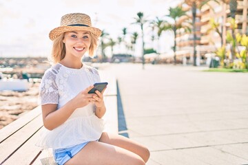Fototapeta premium Young caucasian tourist girl smiling happy using smartphone sitting on bench at promenade.