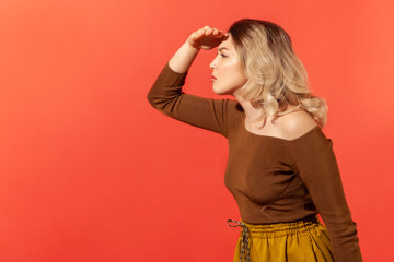 Side view portrait of curious blonde woman holding hand above eyes and peering into distance,...