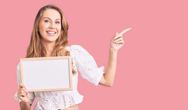 Young caucasian woman with blond hair holding blank empty banner smiling happy pointing with hand and finger to the side
