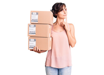 Beautiful young woman with short hair holding delivery packages serious face thinking about...